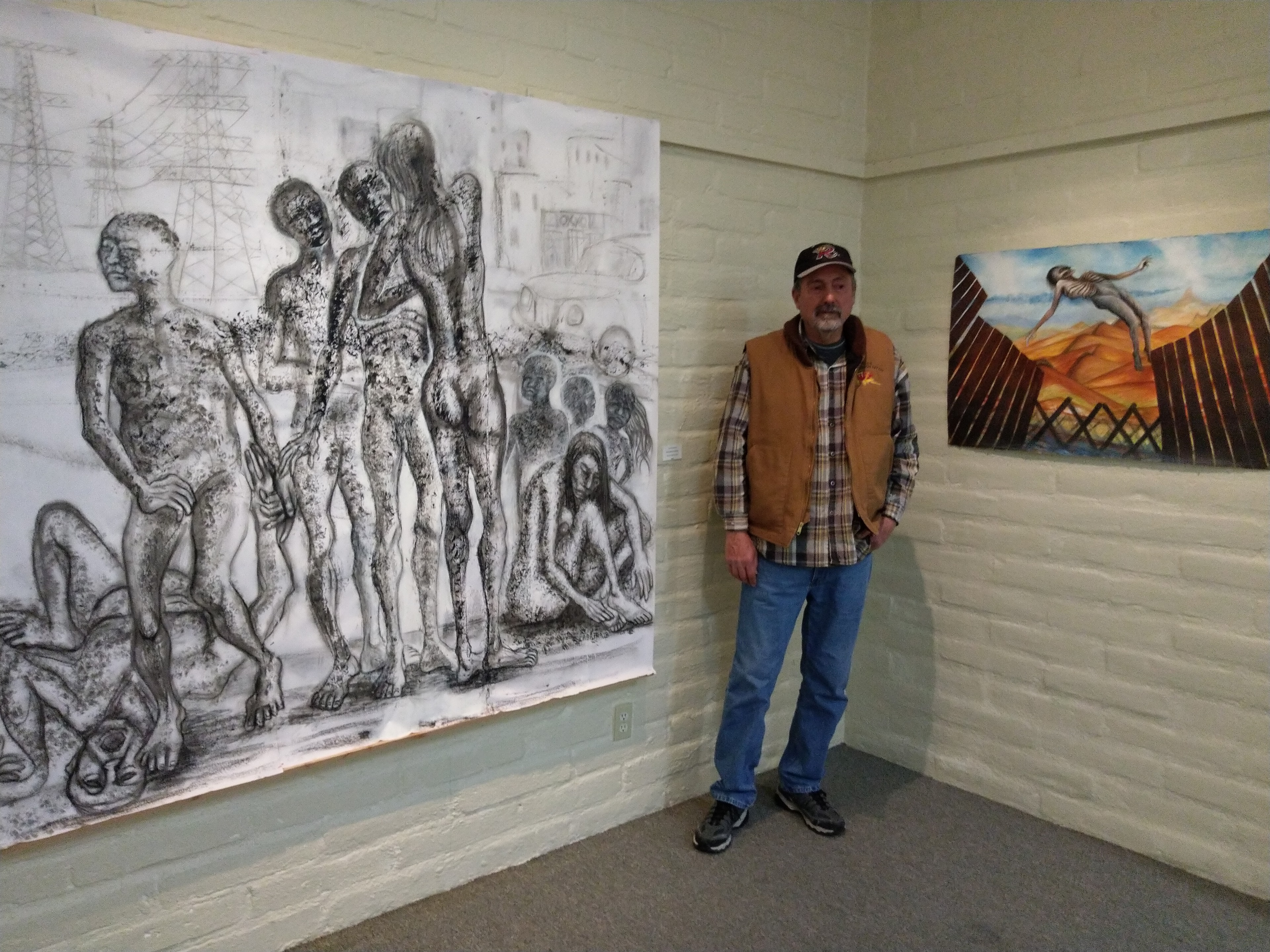 Carlos Cabrera & Ricardo Santos Hernández — Two-Man Exhibition, Hilltop Gallery, Nogales, Arizona. Carlos Cabrera stands before his large-scale drawing — figures rendered in graphite with raw, visceral power, the urban industrial landscape looming behind them. On the wall to the right, Ricardo's painting: a figure arching over the steel bollard wall, the Sonoran Desert burning orange beneath a wide sky. Two artists from Ambos Nogales, two bodies of work, one shared borderlands vision. Carlos Cabrera is co-founder with Ricardo Santos Hernández of the Ambos Nogales Border Art Project. by Ricardo Santos Hernández