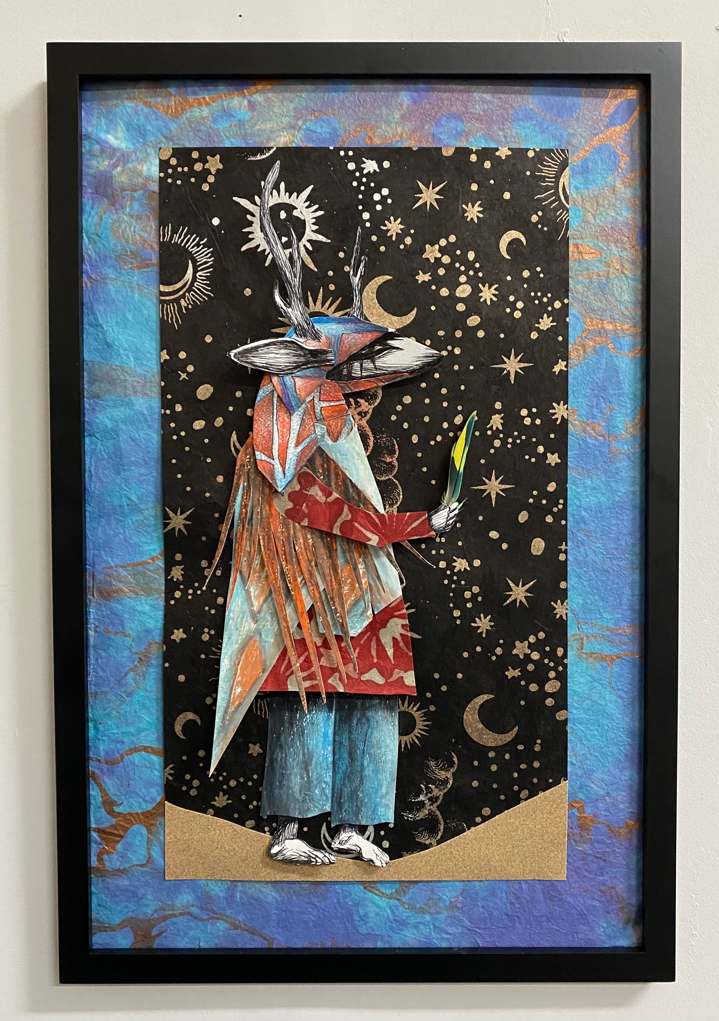 Danzante / Deer Dancer — Ricardo Santos Hernández. Mixed media works on paper. A solitary Yaqui deer dancer stands against a celestial night sky alive with stars, moons, and suns — antlers branching upward like sacred geometry, ceremonial regalia layered in cut paper of red, turquoise, and earth tones, a sacred feather held in hand. The figure stands on desert ground, rooted between earth and cosmos. A meditation on Yaqui spiritual identity, the sacred dance, and the living connection between the people and the land of the Sonora-Arizona desert. by Ricardo Santos Hernández